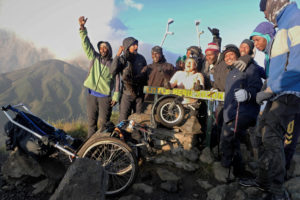 More people are standing on top of a mountain, a woman is in a wheelchair.