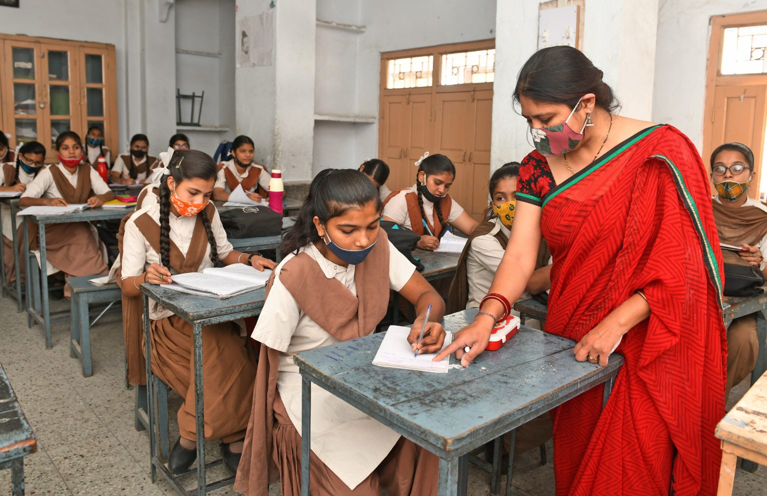 a classroom where several girls sit at school desks, the teacher stands at one of the desks and helps one of the girls who lacks an arm