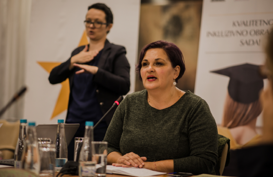 Kvinna med kort brunt hår sitter vid en mikrofon och pratar under ett seminarium