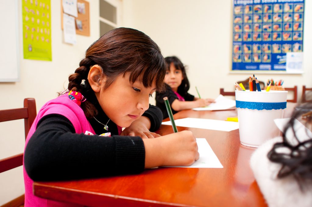 Ung flicka med hörapparat sitter och skriver i skolan hon har håret i fläta och har en rosa väst över svart tröja på sig