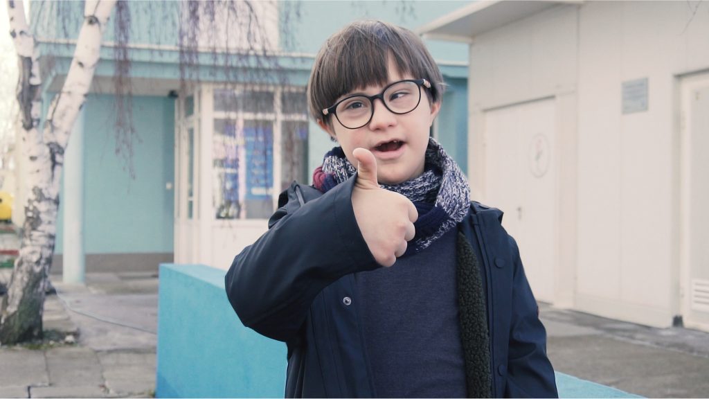 young boy with down syndrome gives thumbs up outside his school he has dark hair, glasses and dark jacket, he smiles big