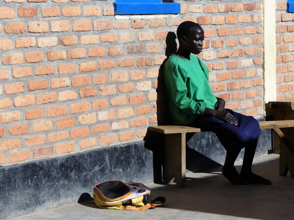 Flicka sitter ensam på en bänk utanför en skolbyggnad i Rwanda hon har blå kjol och grön tröja