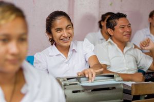 isabell está sentada frente a una máquina de escribir Braille en un salón de clases y sonriendo, tiene un uniforme escolar blanco y cabello oscuro recogido en una borla
