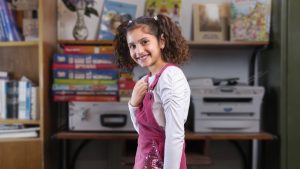 Mitra har mörkt hår uppsatt i två toffsar hon har vit tröja och rosa hängselbyxor hon ler och har ena handen över bröstet hon står i ett klassrum