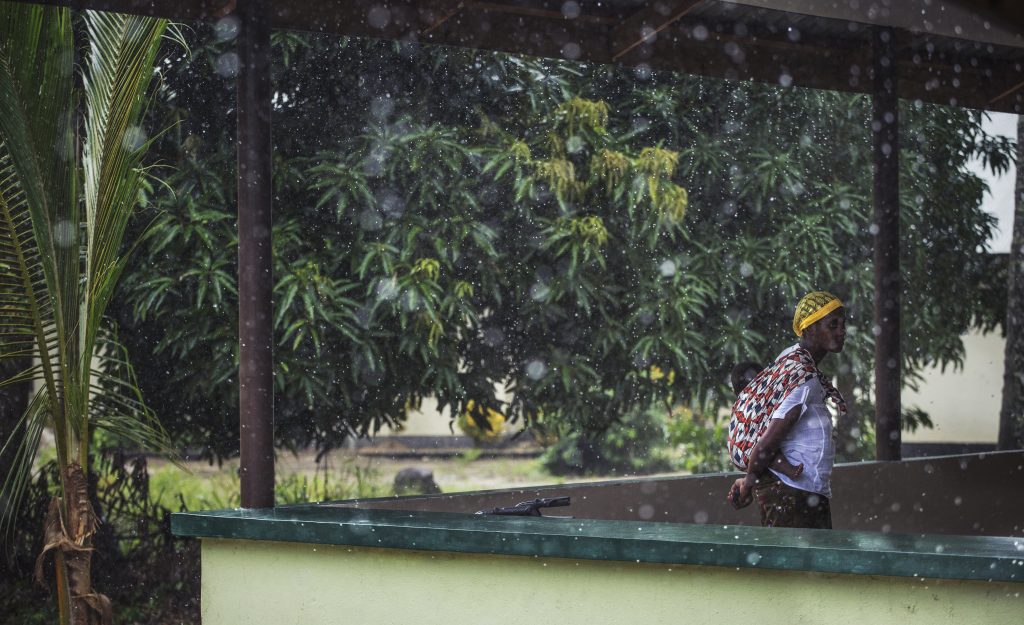una mujer está parada en un balcón con su pequeño hijo en la espalda, hay árboles y lluvia en el fondo