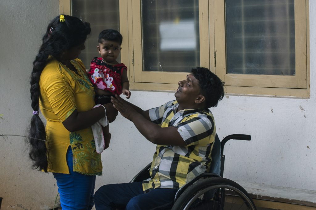 un hombre se sienta en una silla de ruedas y extiende sus brazos hacia su pequeña hija que está sentada en los brazos de su madre