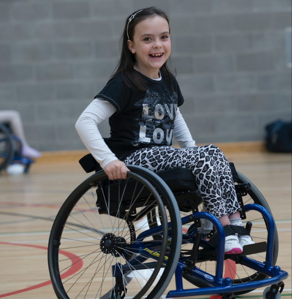 una niña está sentada en una silla de ruedas en un gimnasio, sonríe a lo grande y tiene el pelo castaño