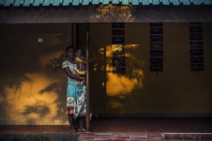 A woman stands outside a house with a child in her arms.
