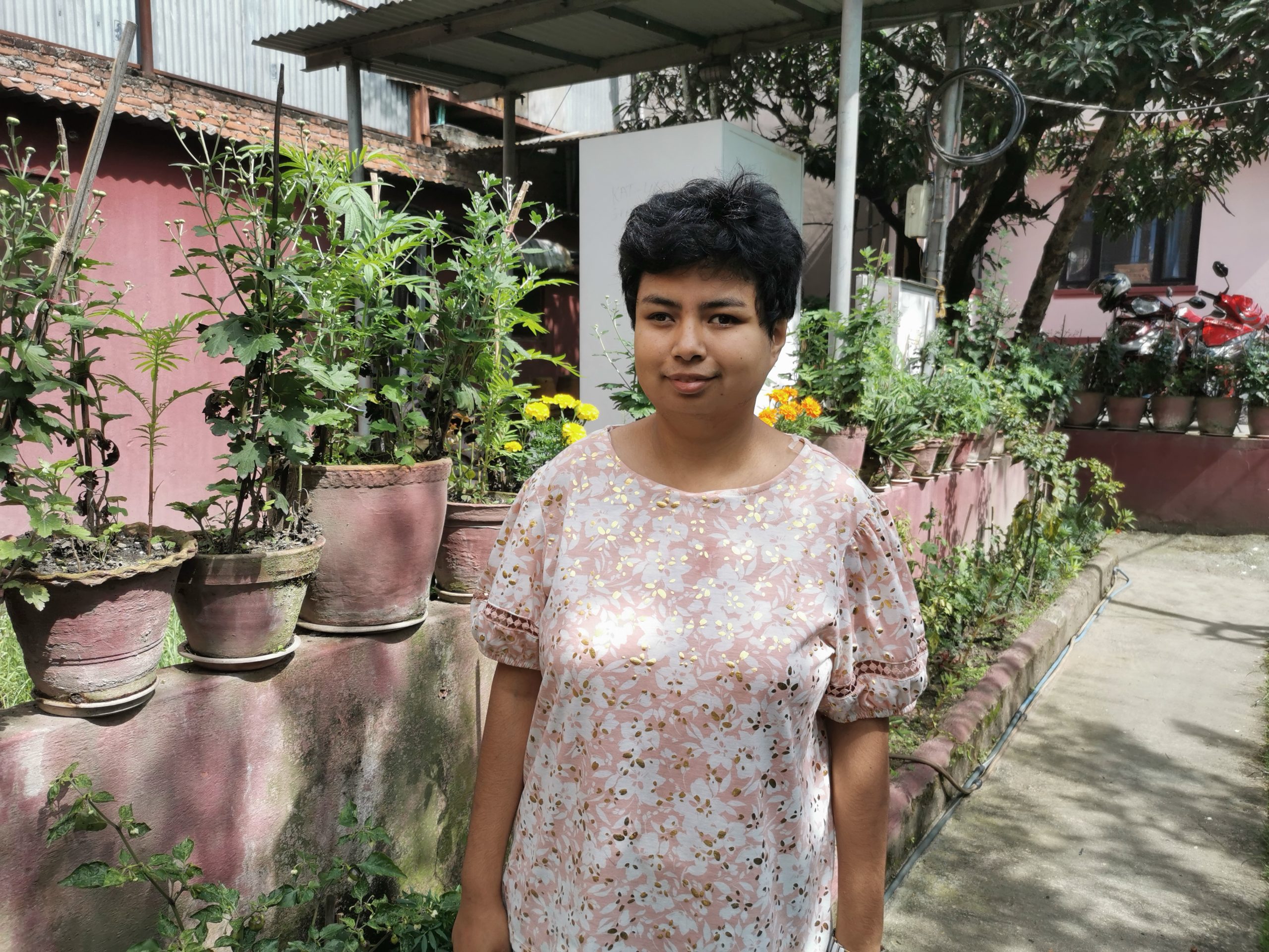 Ayushma está de pie en un jardín con plantas en macetas verdes detrás de ella, lleva un suéter rosa y el pelo corto y negro.