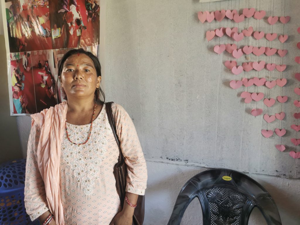 Indira nosi ružičastu bluzu i crnu kosu, stoji uza zid u ćerkinoj učionici. na zidu okače roze papirna srca koja su djeca izrezala.