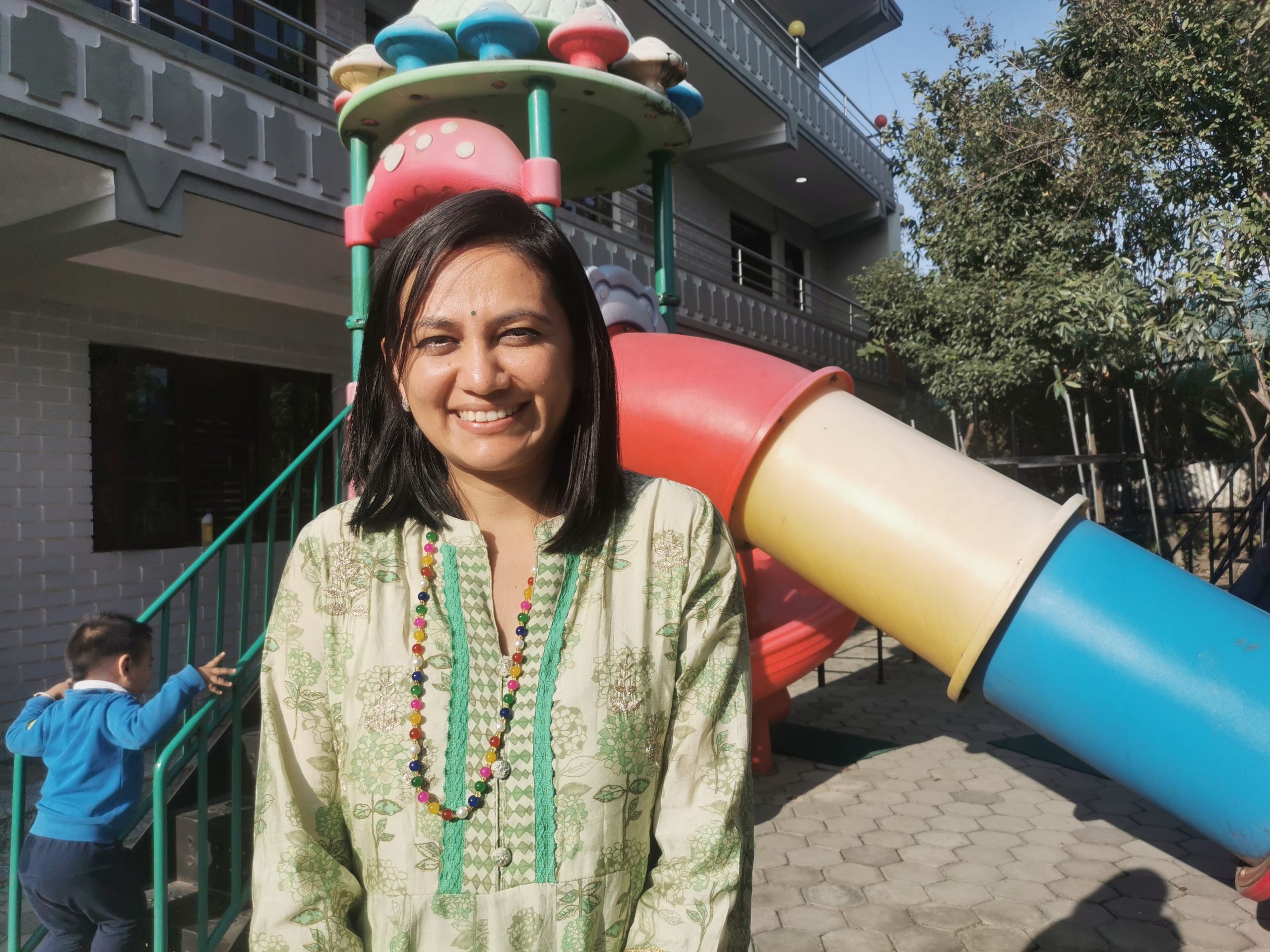 Sangita has black shoulder length hair, she smiles into the camera. behind her, a small child is climbing a climbing frame.