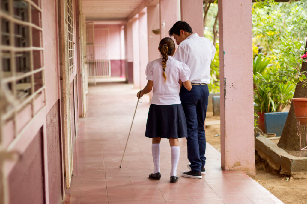 En flicka i skolkläder och vit käpp går i en korridor och blir ledsagen av en skolkamrat.