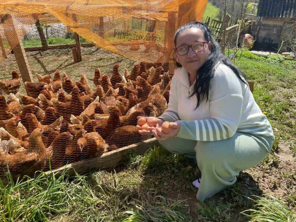 Muhida sitter på huk framför ett hönshusö Hon har mörkt långt hår och glasögon.