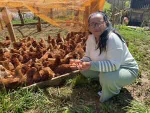 Muhida sitter på huk framför ett hönshusö Hon har mörkt långt hår och glasögon.