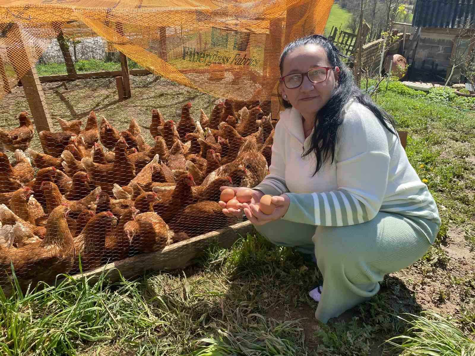 Muhida sitter på huk framför ett hönshusö Hon har mörkt långt hår och glasögon.