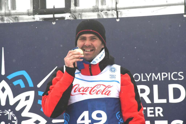 Rasim står utomhus och har en sportväst på sig och mössa. Han biter glatt i en guldmedalj.