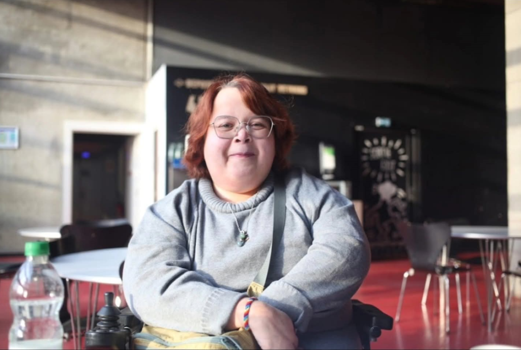 Nora har kort mörkt hår och glasögon. Nora sitter i rullstol.